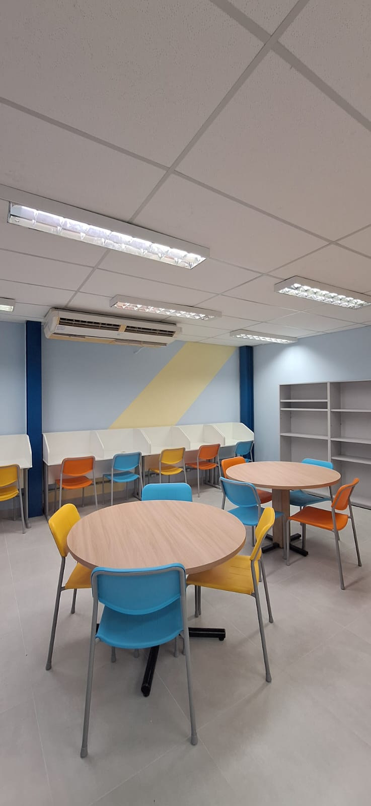 Espaço de leitura do Colégio Anglo com mesas redondas, cadeiras coloridas e estantes.